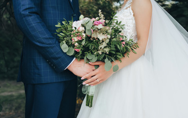 The bride in a white wedding dress and the groom in a blue checkered suit stand against each other and hold a large bouquet of white and pink roses with green foliage.
