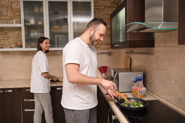 Happy young couple preparing breakfast, man fries broccoli, scrambled eggs. Girl slices fresh vegetables. On self-isolation during the coronovirus pandemic covid-19.
