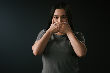 Front view of beautiful plus size girl gesturing silence by covering mouth with hands on black background