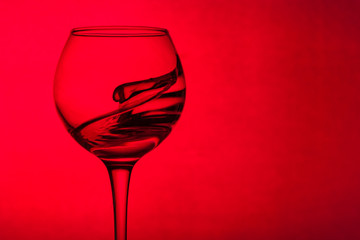 Wine glass with liquid on a red background. The liquid in the glass splashes. Copy space