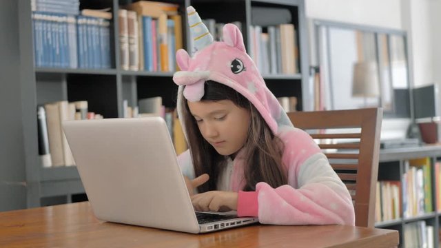 ni&ntilde;a de melena con capucha de unicornio rosa estudia en un port&aacute;til junto a una librer&iacute;a