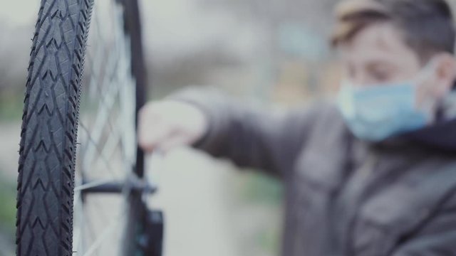 A 13 year old teenager is repairing his bike with a medical mask on his face. The concept of changing the ordinary life of people in connection with the coronavirus pandemic.