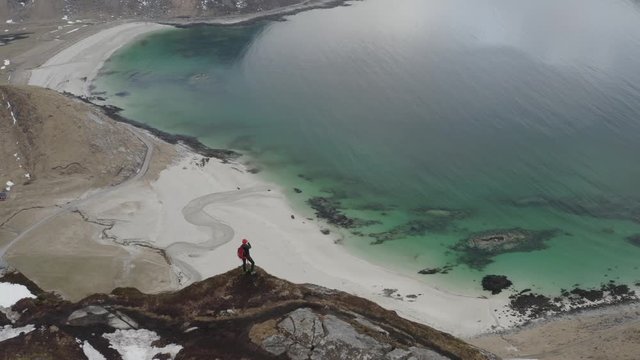 aerial video from drone. woman standing on the edge of mountain above a beach and taking photos of incredible landscape. located in lofoten islands north of norway.