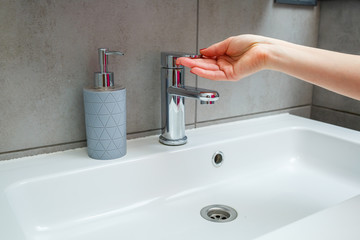 White sink with a silver faucet in the bathroom. Gray can with liquid soap for hands. Turning on tap water, personal hand hygiene