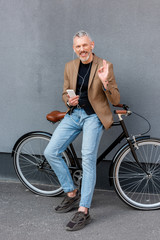 Plakat cheerful businessman in earphones listening music while showing ok sign and holding smartphone near bicycle outside