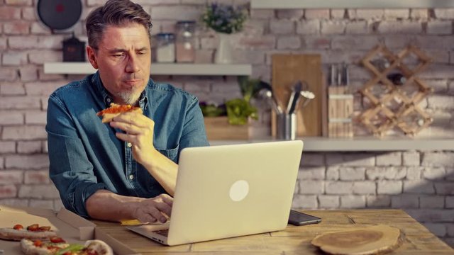 Man working from home on laptop computer, sitting at table in kitchen, eating online ordered pizza.