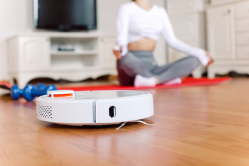 Woman sitting in lotus pose doing yoga at home while autonomous vacuum cleaner cleans the floor....
