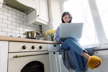 Portrait of woman working on laptop while cooking in kitchen. Comfortable light space to work at home 