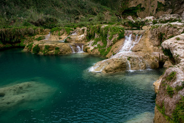 Poza de agua color turquesa con formaciones de piedra blanca caliza debajo del agua rodeado de caídas de agua en la roca cubierta de helechos 