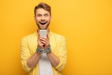 Cheerful man with metal chain around hands holding smartphone on yellow background