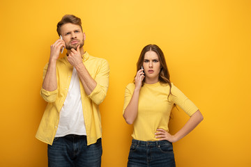 Confused couple talking on smartphones on yellow background