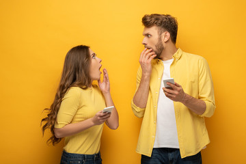 Surprised couple with smartphones looking at each other on yellow background