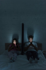 Naklejka premium Young couple with smartphones sitting near model of smartphone on bed at night