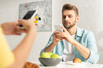 Selective focus of man using smartphone near girlfriend during breakfast in kitchen