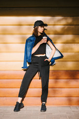 Beautiful stylish brunette girl with coffee in a black cap and denim jacket outdoors vertical photo