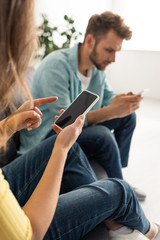 Selective focus of woman pointing with finger at smartphone with blank screen near boyfriend on couch