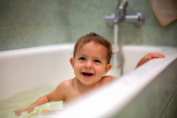 Cute Caucasian baby taking a bath, smile and look up, resting on the side of the bath. water splashes, in the background a green bathroom in blur. close-up, soft focus