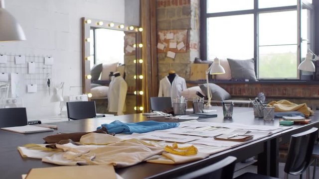 Zoom out of tailor worktable with fabric, fashion sketches and sewing supplies in studio with loft interior