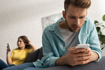 Selective focus of addicted man using smartphone near girlfriend on sofa