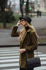 Girl in the coat and black hat standing on the crosswalk and looking ahead holding her glasses