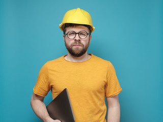 Suspicious male foreman or engineer in a construction helmet and round glasses stands with a laptop and looks with squinted eyes.