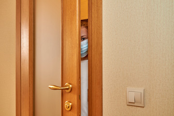 Obraz premium A little scared girl at medical protective face mask is looking through the door slit. A girl is watching someone or something through ajar door. Coronavirus or Covid-19 concept.