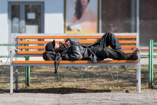 A Poor Homeless Man Is Lying On A Bench And Sleeping In Cold Weather.