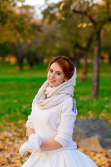 Cute girl in long white wedding dress posing in rural path among autumnal trees in forest in golden hour atmosphere.