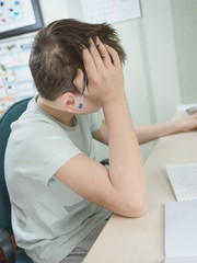 A teenager boy sits at a table in his room, doing homework. He's bored.