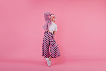 Full-length portrait of adorable european woman wears pink striped pants and fur jacket during photoshoot. Refined girl with short colorful hair posing in trendy clothes in studio.