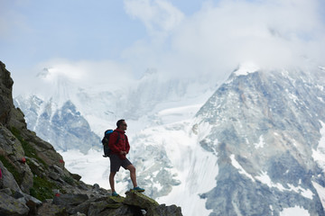 Side view of tourist with backpack standing on big stone, looking at beautiful mountains with snow. Trekking, mountain hiking, man reaching peak. Wild nature with amazing views. Sport tourism in Alps.