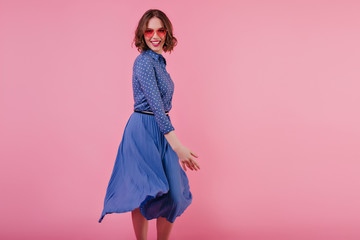 Slim caucasian woman in vintage skirt posing on pink background and smiling. Cheerful european girl in trendy blouse and sunglasses having fun in studio.