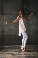 Beautiful young slender blond woman with curly hair in white trousers, a blouse and shoes in a loft studio is dancing. Soft selective focus.