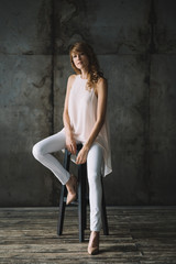 Fototapeta premium Beautiful young slender blond woman with curly hair in white trousers, a blouse and shoes in a loft studio is sitting on a bar stool. Soft selective focus.