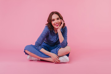 Studio portrait of amazing european girl in trendy blouse sitting on pink background. Beautiful white female model in skirt posing on the floor with legs folded.