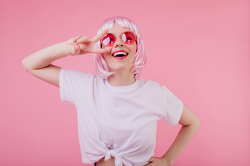 Ecstatic caucasian girl in trendy white t-shirt posing with peace sign and laughing. Indoor photo of dreamy european woman in shiny peruke and sunglasses.