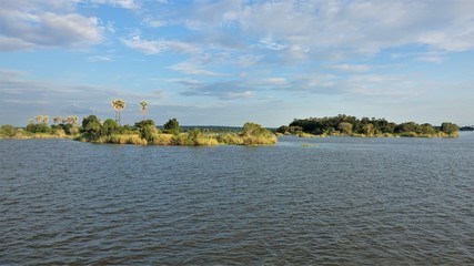 The Zambezi River flows calmly. On it are small islands covered with grass, bushes, silhouettes of palm trees are visible. There are beautiful clouds on the blue sky.