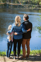 Rear view of loving family of mother, father and little daughter standing in embrace near lake in countryside and looking at scenic view