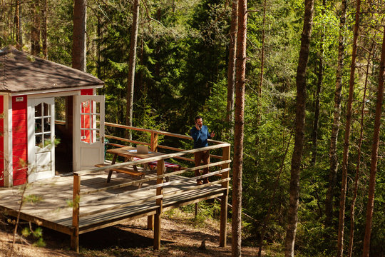 From Distance Shot Of Middle Aged Man Talking On Cell Phone Standing On Terrace Of Country House Surrounded By Pine Trees, Open Laptop On Wooden Table Behind Him