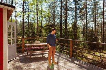Wide shot of middle aged man texting on cell phone standing on porch of country house surrounded by pine trees, open laptop on wooden table behind him