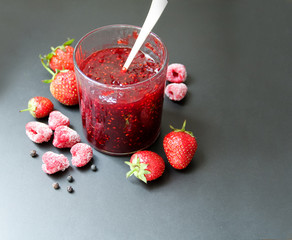 Strawberry and raspberry jam with black pepper in a glass with a silver spoon. Dark background. Back view.