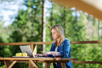 Side view of young woman working on laptop computer sitting at wooden table on balcony of...