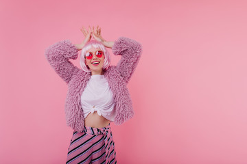 Pleased young woman in trendy outfit making funny faces during photoshoot in peruke. Studio photo of laughing girl in fur jacket and sunglasses.