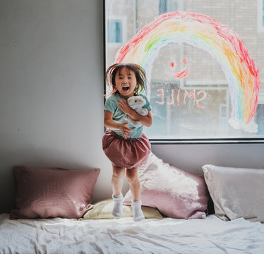 虹とスマイルのメッセージが描かれた窓の前　ベッドの上で飛び跳ねてはしゃぐ元気な女の子
