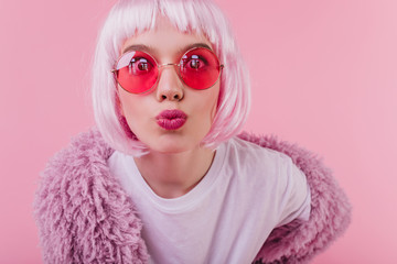 Close-up shot of surprised young lady in peruke posing with kissing face expression in studio with pink interior. Jocund caucasian girl in periwig enjoying photoshoot.