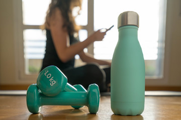 Girl doing gymnastics and looking at her mobile phone. Close-up of green weights and bottle
