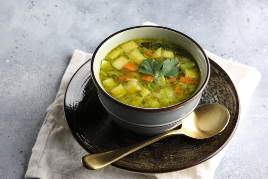Lean vegetable soup with potatoes, zucchini, cabbage, carrots and herbs in a deep bowl with a spoon on a light grey background. Fresh parsley. Background image, copy space