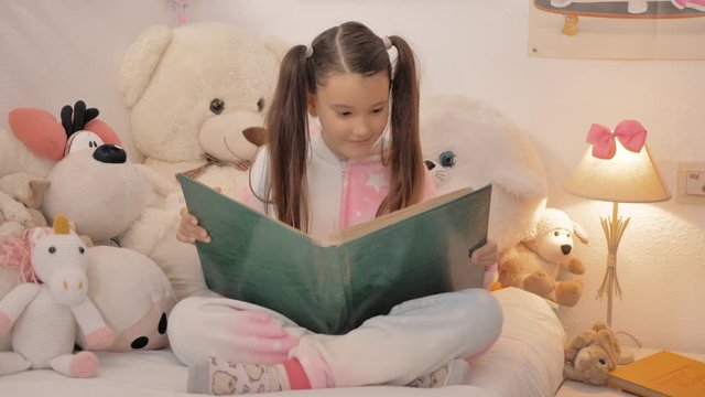 joven peque&ntilde;a con coletas largas mura &aacute;lbum de fotos en papel sentada en la cama