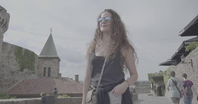 Young woman walking in the city, ancient wall background. Beautiful fashion model face with sunglasses, travel and explore the old town fortress in Belgrade Serbia, s-log ungraded