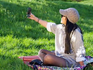 Beautiful Asian woman wearing a hat and sunglasses having a relaxing time in the city park sitting on a colorful blanket surrounded with a green grass using her phone making a selfie.